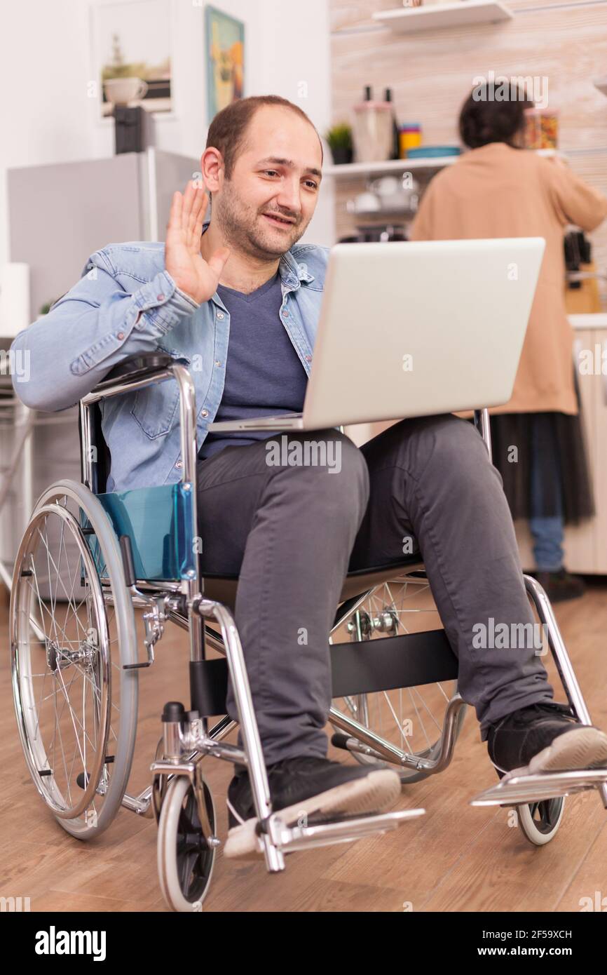 Disabled entrepreneur in wheelchair waving during a video call on laptop while wife is cooking lunch. Disabled paralyzed handicapped man with walking disability integrating after an accident. Stock Photo