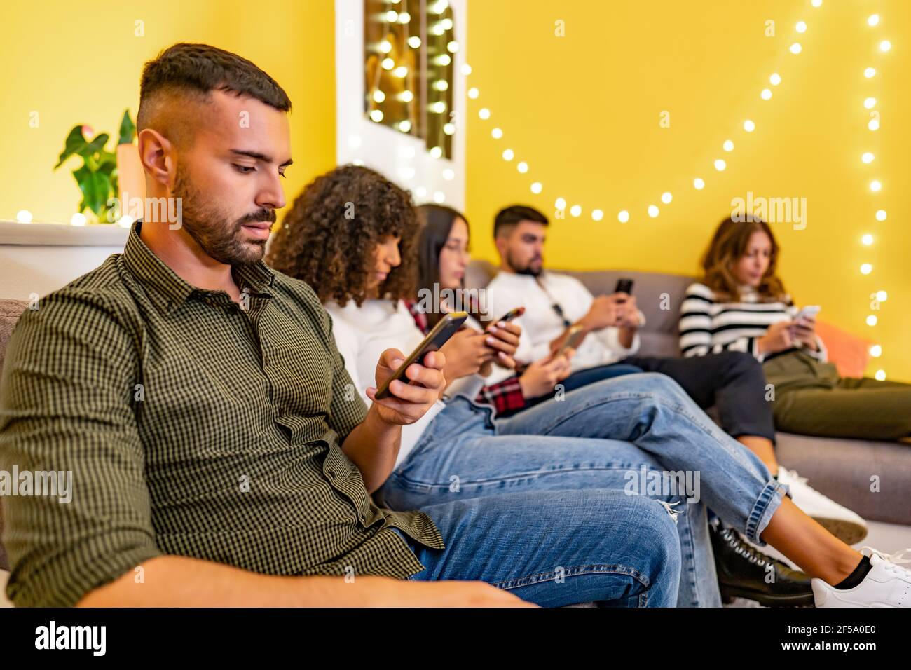 Group of serious young student friends at home sitting on the couch spend time using smartphones without real interaction between them. New social and Stock Photo