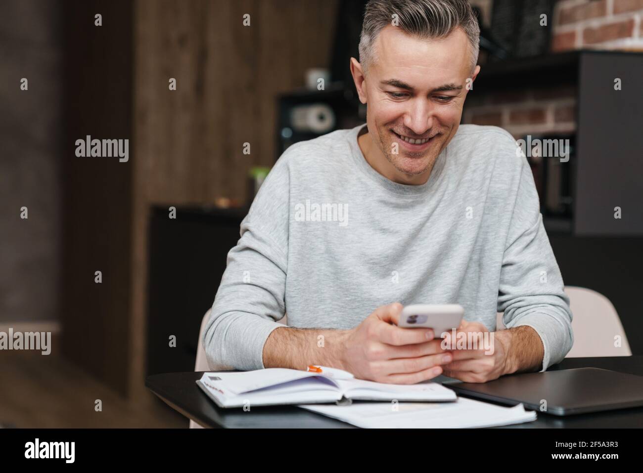 Cheerful handsome man smiling and using mobile phone while working at home Stock Photo