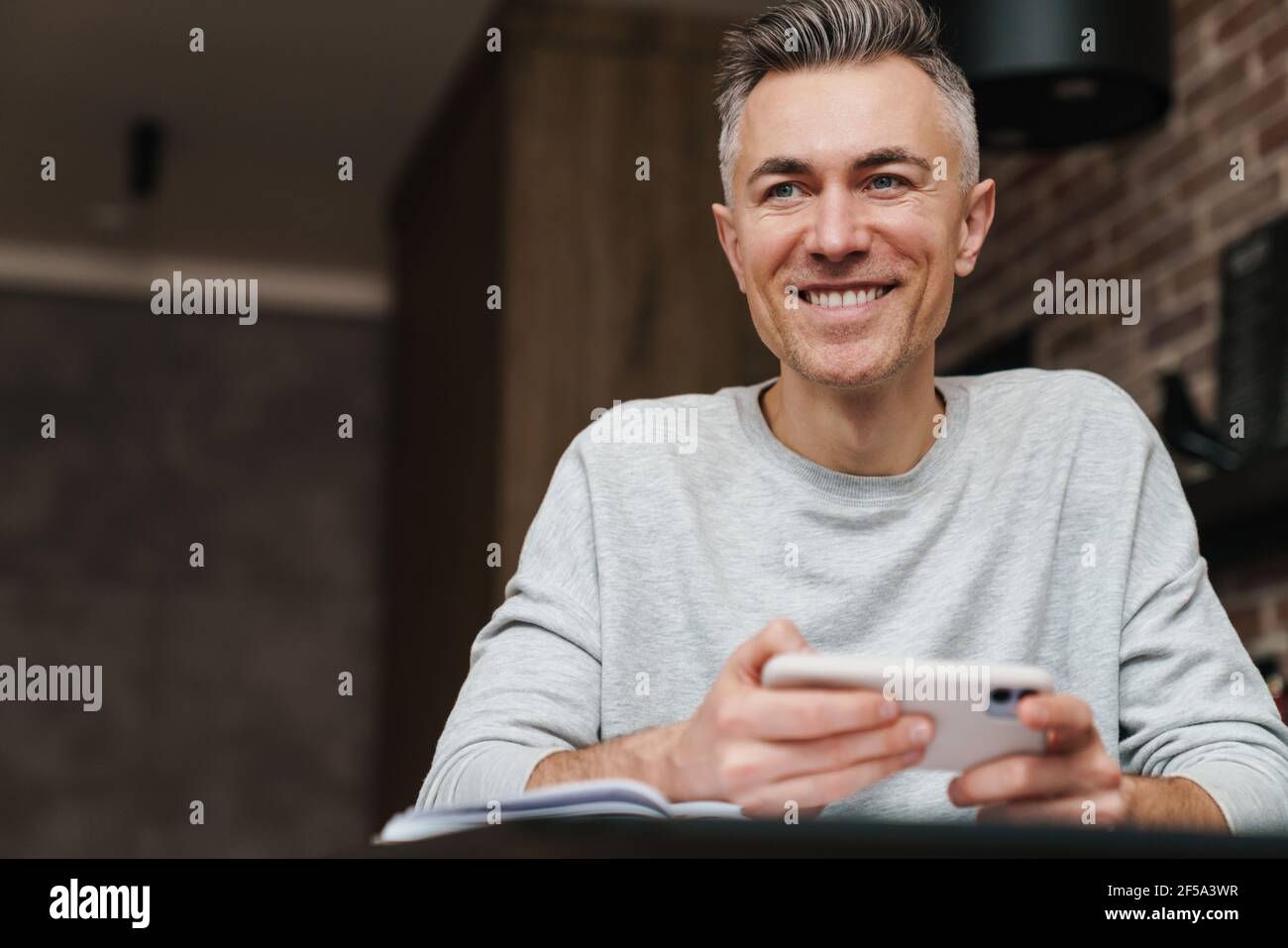 Cheerful handsome man smiling and using mobile phone while working at home Stock Photo