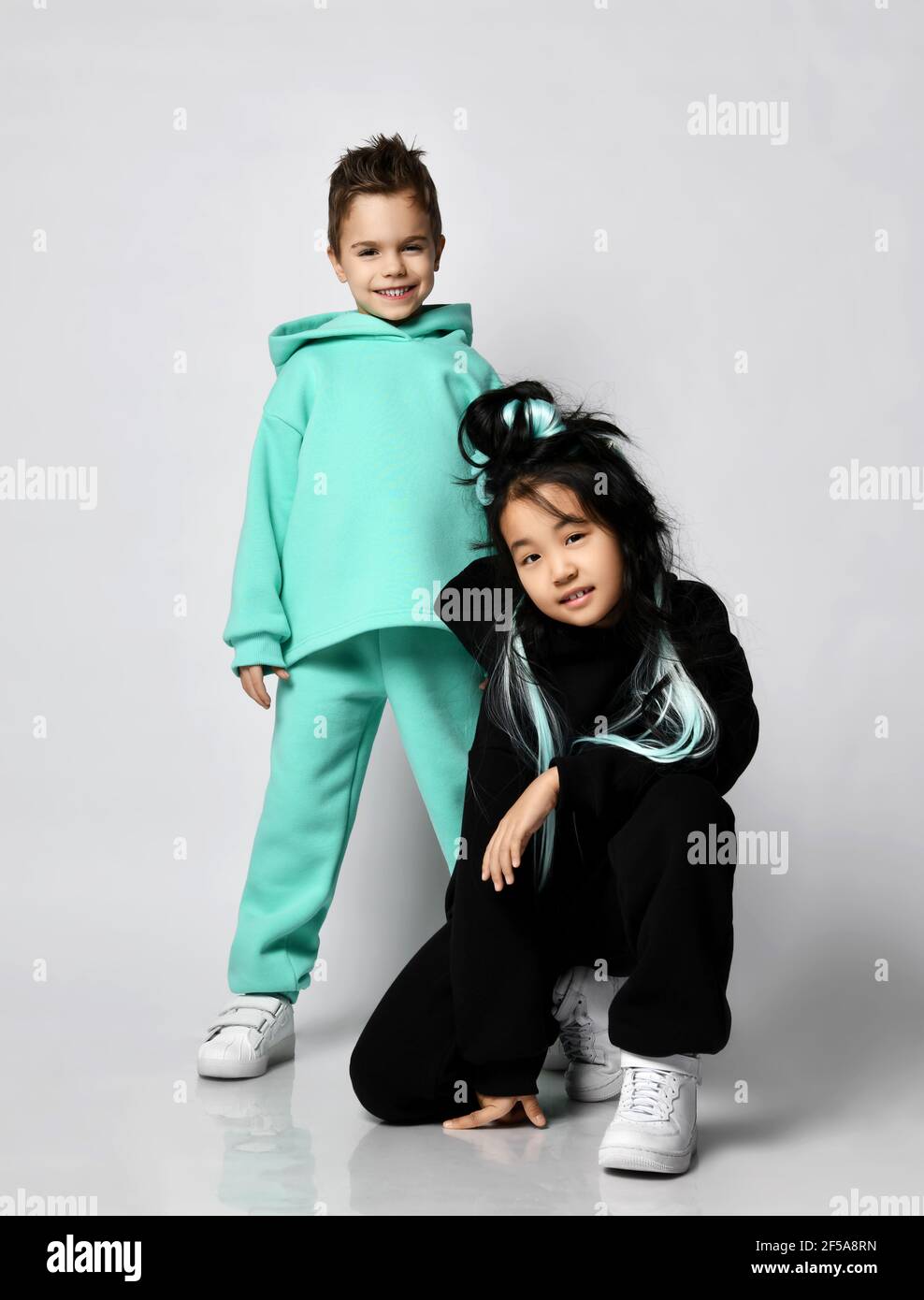 Friends at playground. Kid girl in black hoodie and pants sits squatted at kid boy standing in green, mint sports suit Stock Photo