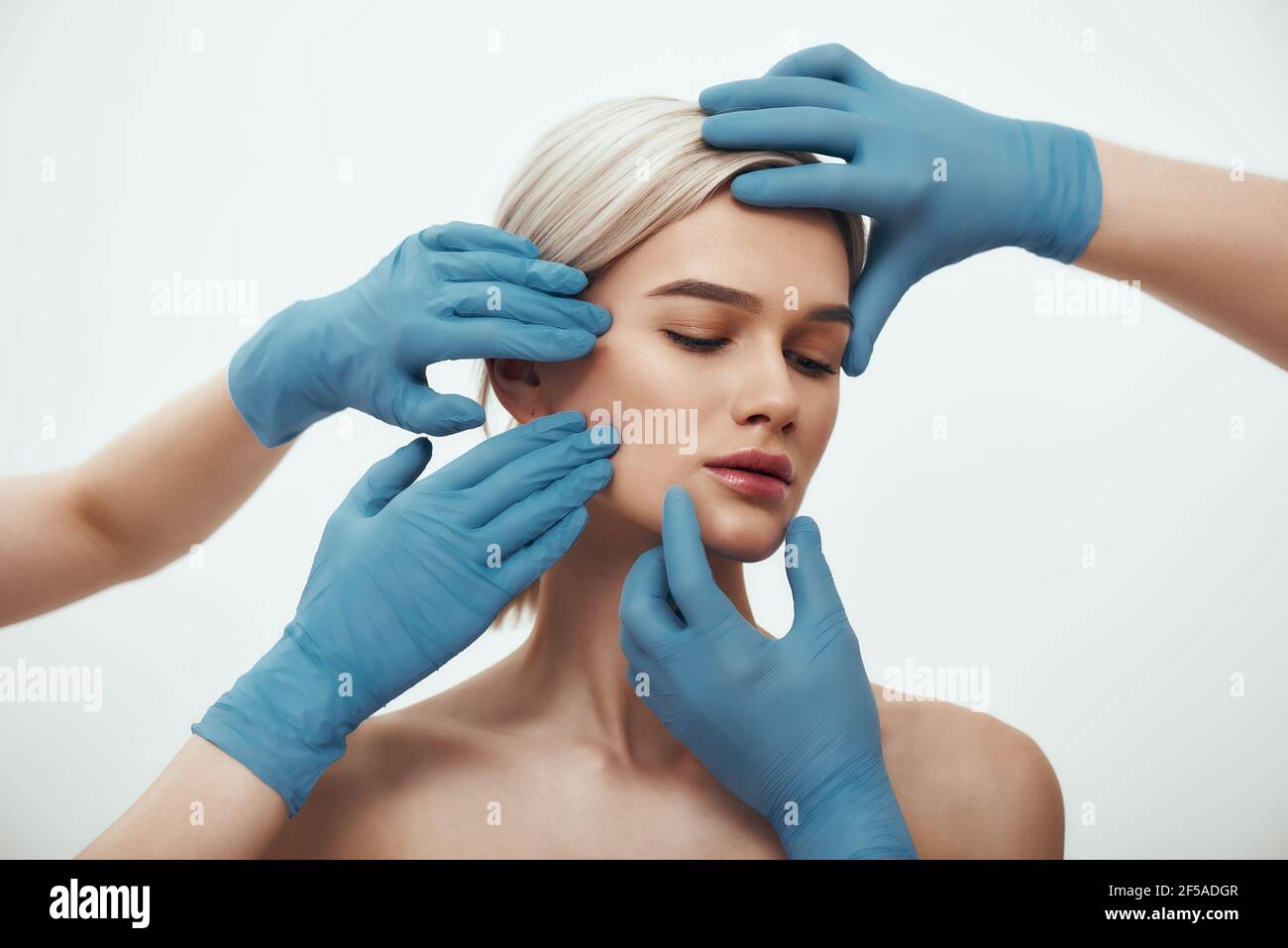 Before facial surgery. Beautiful blonde woman keeping eyes closed while doctors in blue medical gloves examining her face Stock Photo