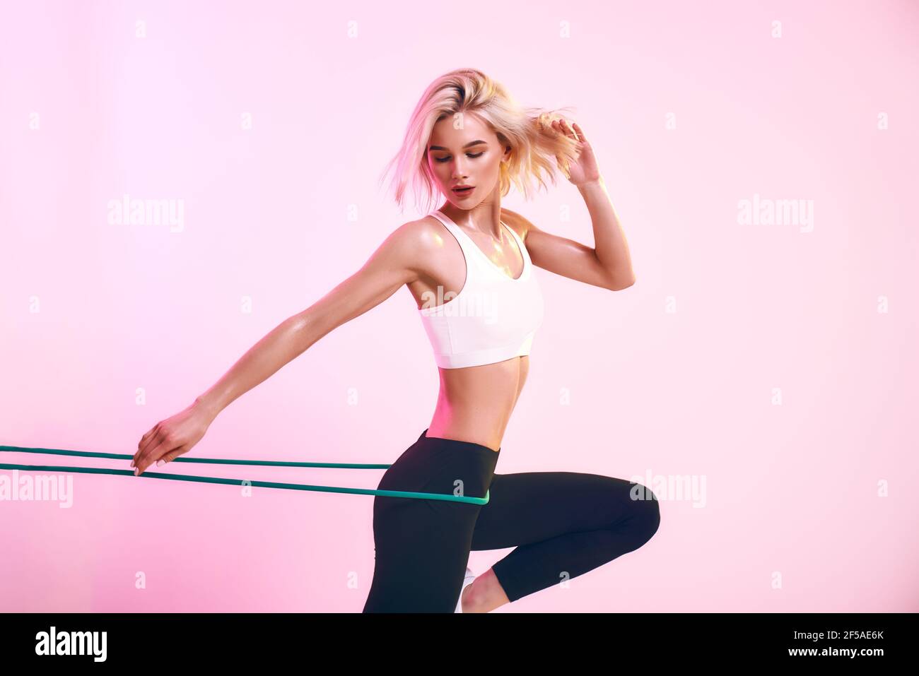 Fitness girl. Sporty beautiful woman in white top and black leggings exercising with resistance band while standing against pink background in studio Stock Photo