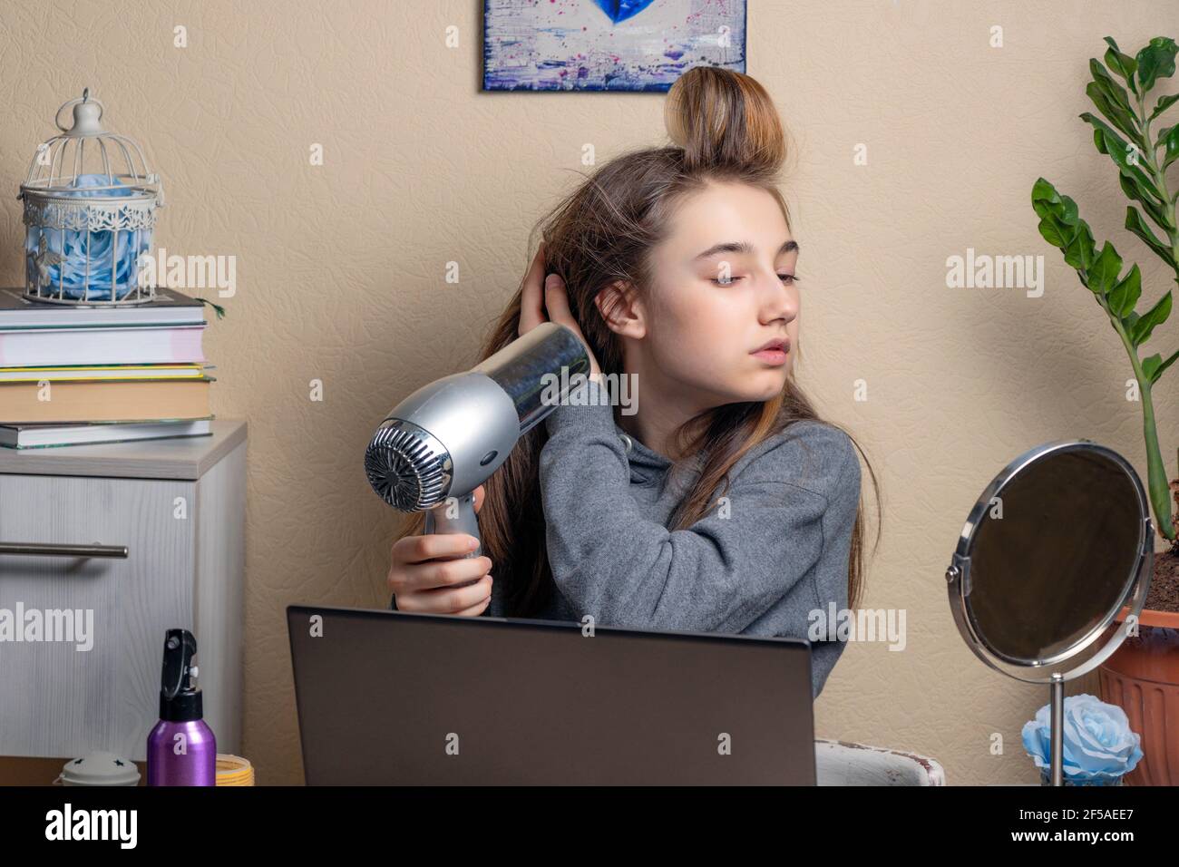 beautiful teenage girl doing her hair in front of laptops. A teenage girl with curlers and a hairdryer looks at the laptop screen. Online beauty conce Stock Photo