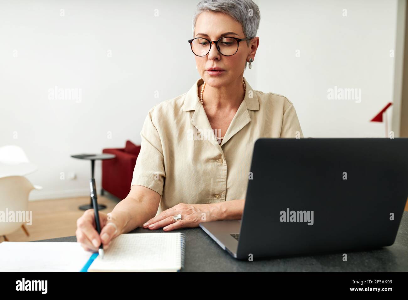 Mature woman writing on a note pad while using laptop computer at home Stock Photo