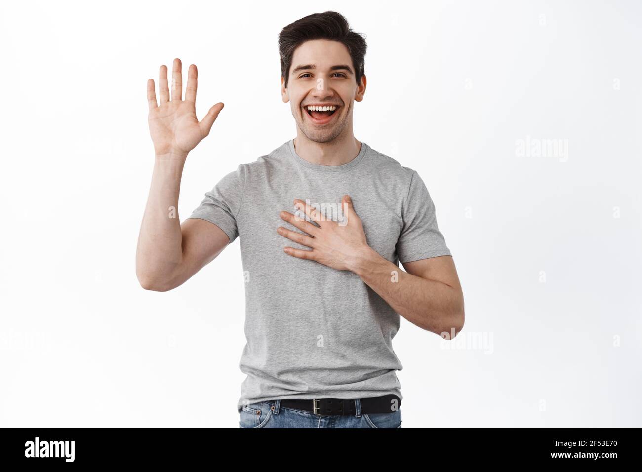 Happy smiling adult man introduce himself, raising hand and hold palm on chest, swearing, making an oath or promise, standing cheerful against white Stock Photo