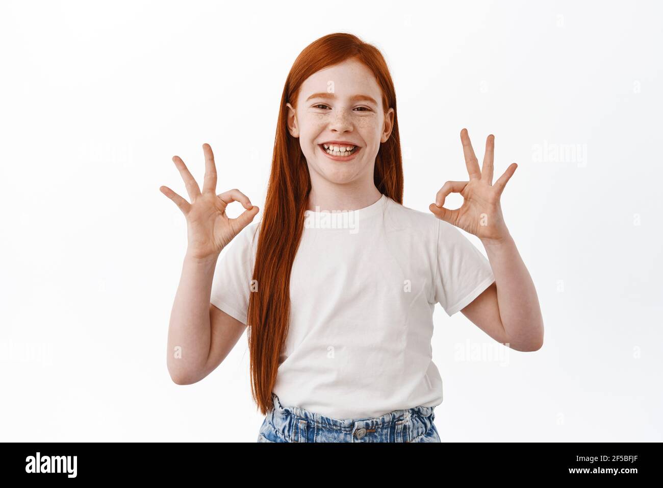 Happy little girl with ginger hair shows okay signs and smiles satisfied. Pleased young child with red head say yes, like and approves, praise good Stock Photo