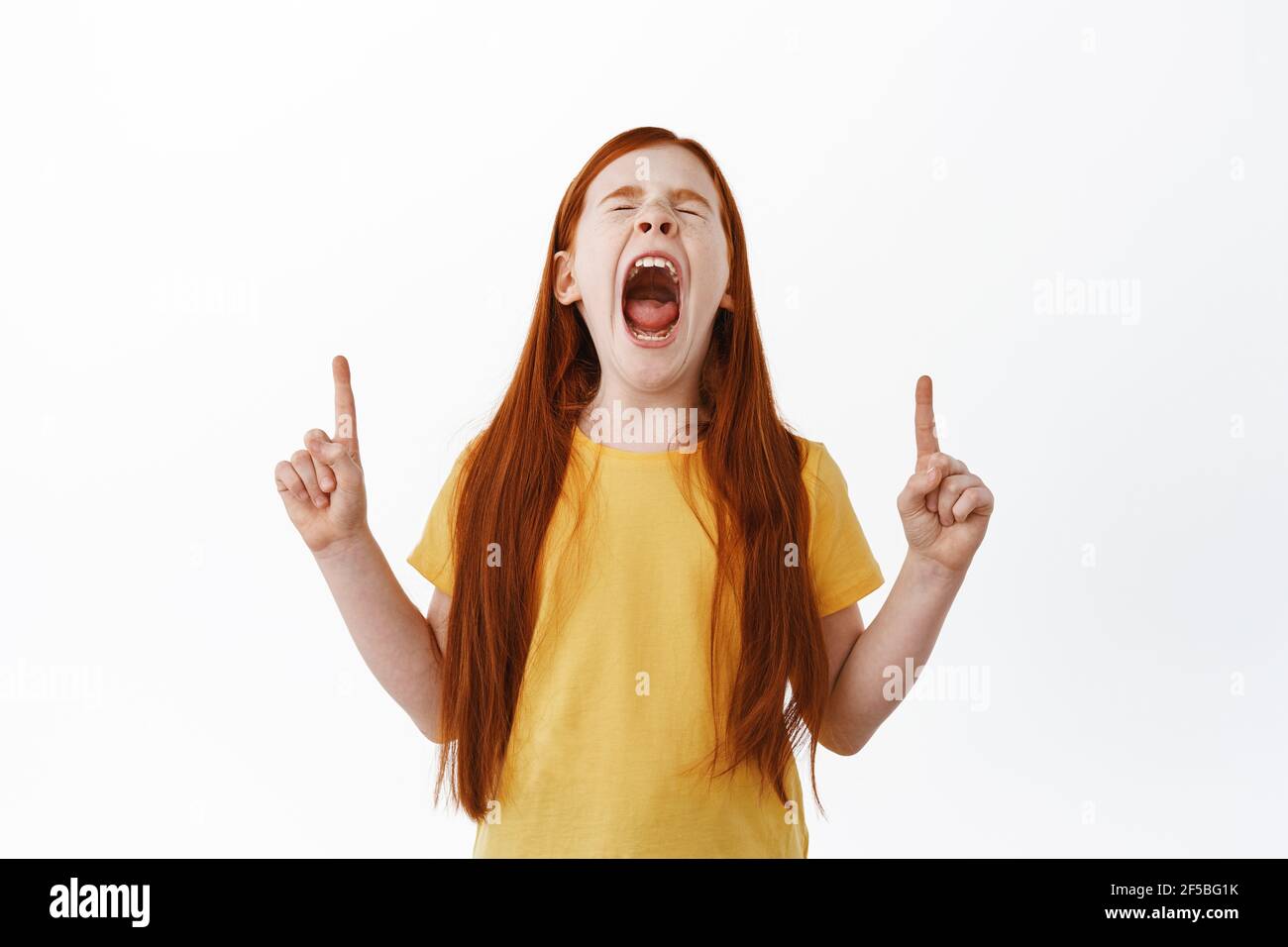 Little red haired girl screaming and pointing fingers up. Ginger kid with freckles shouting and showing something aside on copy space, white Stock Photo