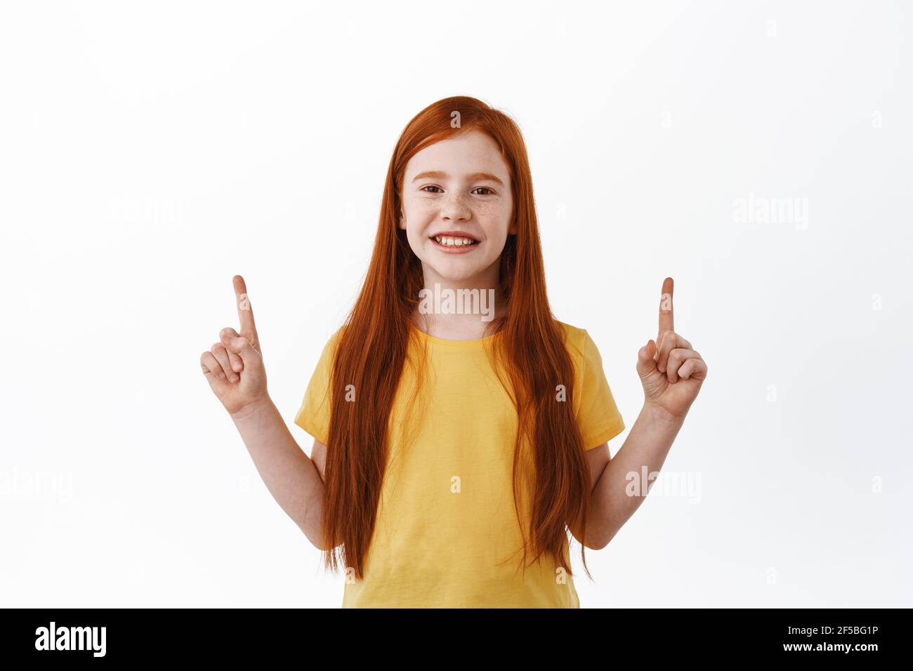 Adorable red-haired girl, little kid with long ginger hair pointing fingers up and smiling pleased, shows something, promotional text on copy space Stock Photo