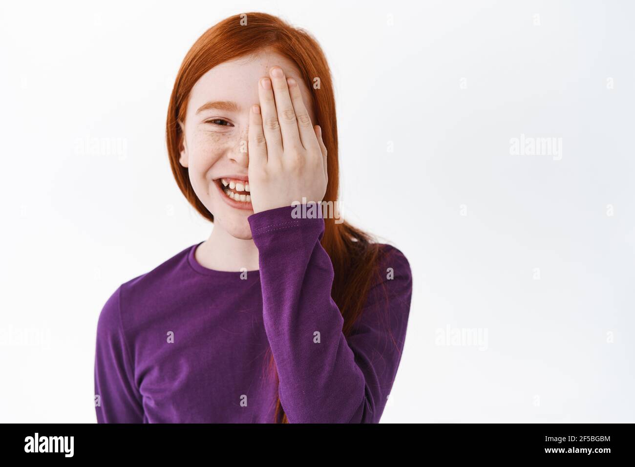 Happy redhead little girl, ginger kid hiding half of face behind hand