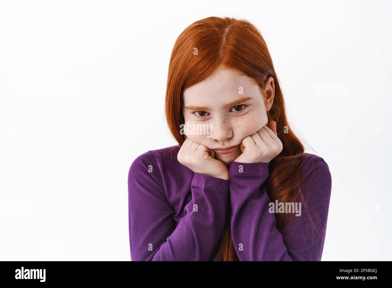 Portrait of cute redhead girl pouting, making coy sulking face and look gloomy from under forehead, standing upset against white background Stock Photo