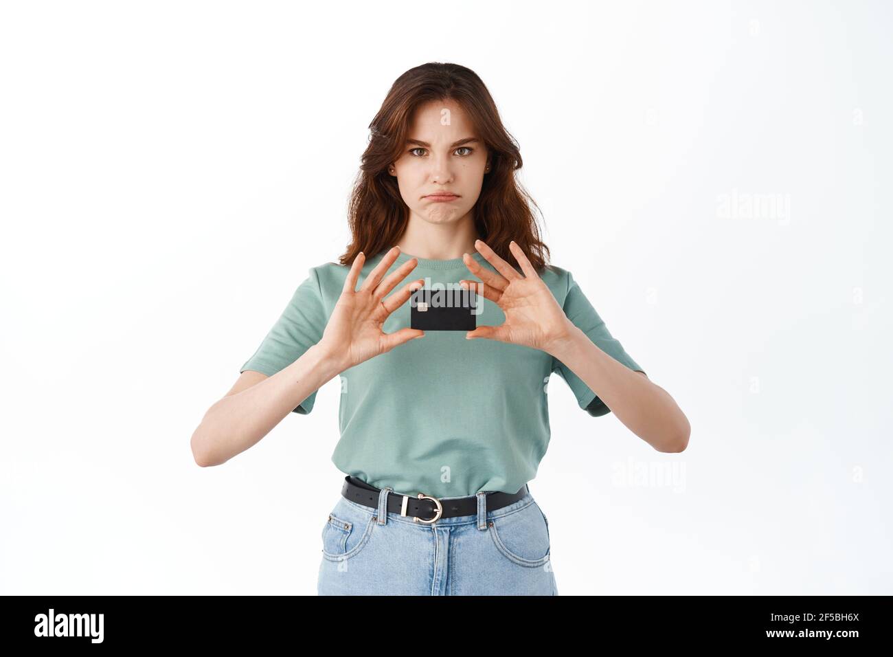 Sad young woman in t-shirt showing her plastic credit card and grimacing upset, having bad mood, standing disappointed against white background Stock Photo