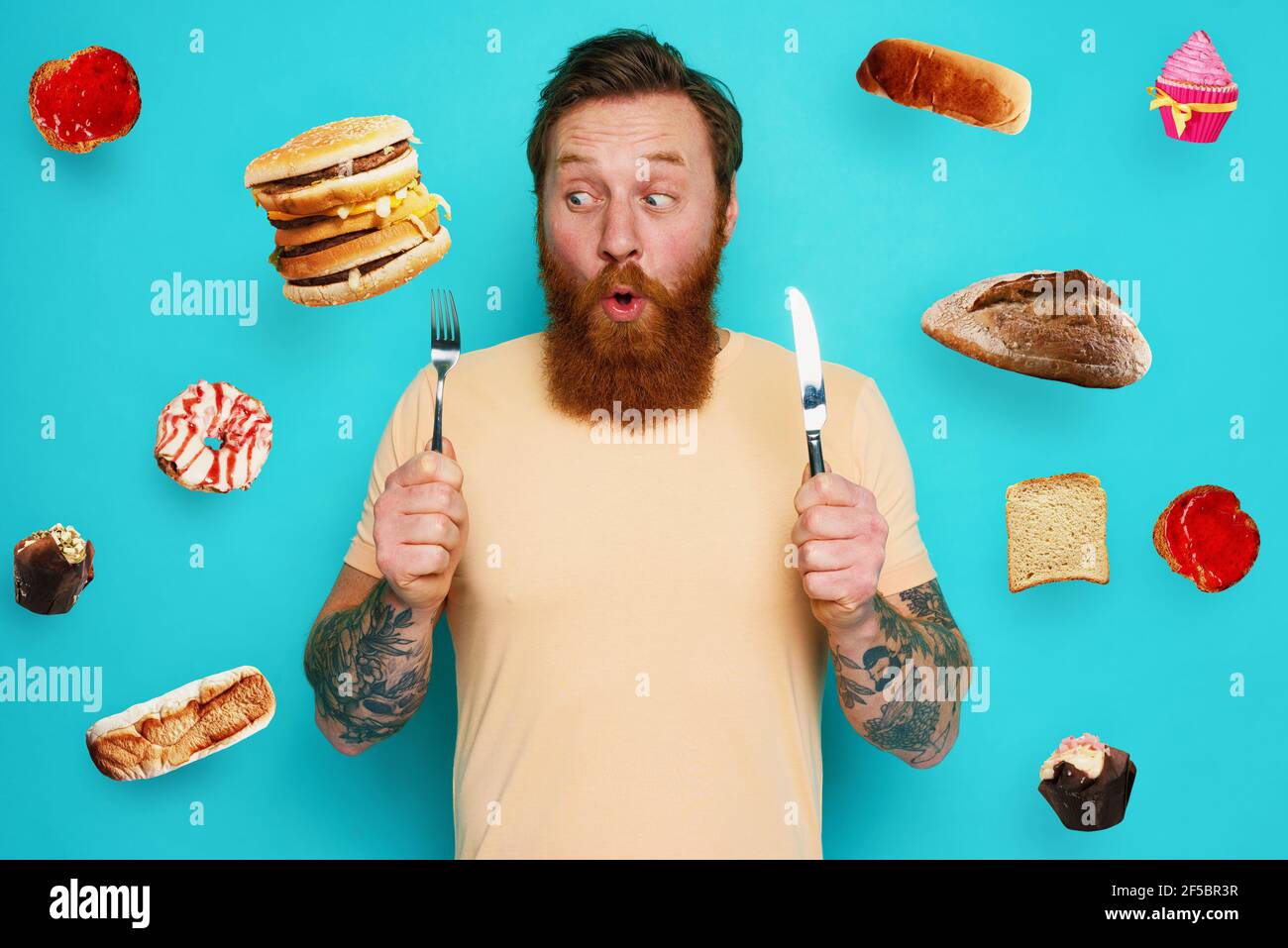 Man with tattoos is ready to eat sandwich and sweets with cutlery in hand. cyan background Stock Photo