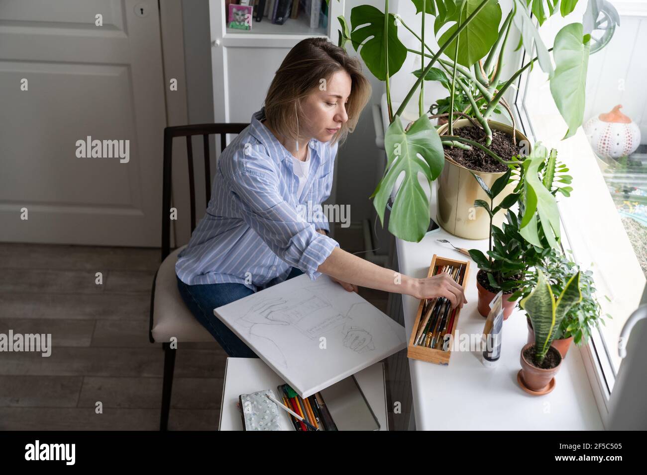 Woman artist paints a picture on canvas, makes pencil sketches, sitting by the window. Slow living. Stock Photo