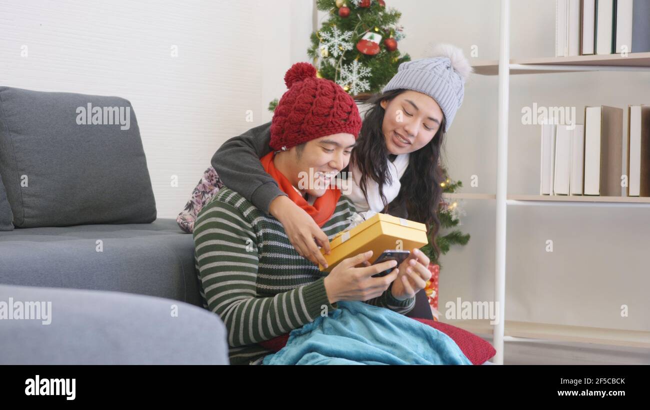 Happy Asian beautiful young family couple husband and wife in home living room, woman hide surprise giving gift box her boyfriend present during play Stock Photo