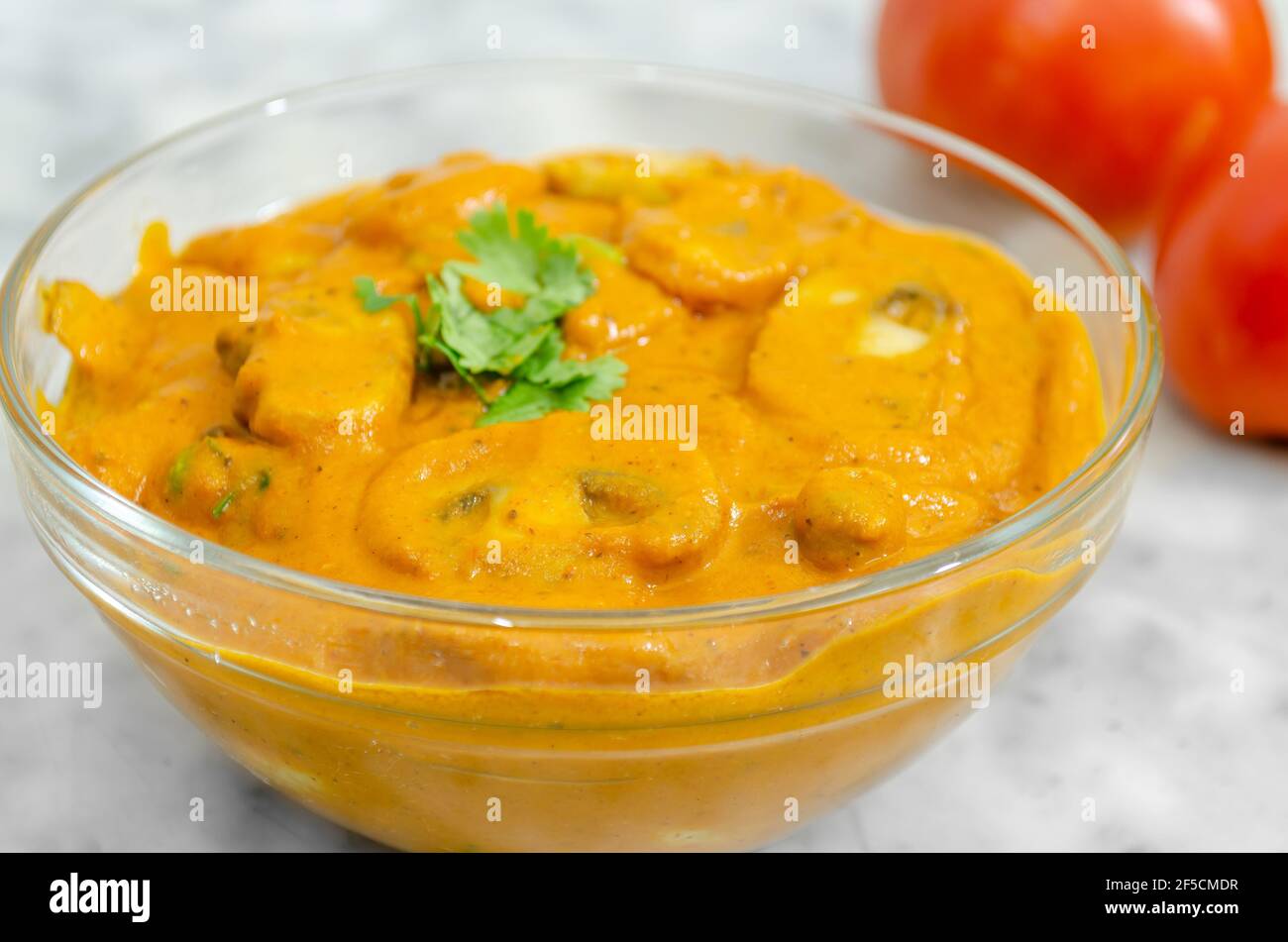 Closeup of A very tasty Mushroom Masala in a glass bowl garnished with coriander leaves and red tomatoes in the background Stock Photo