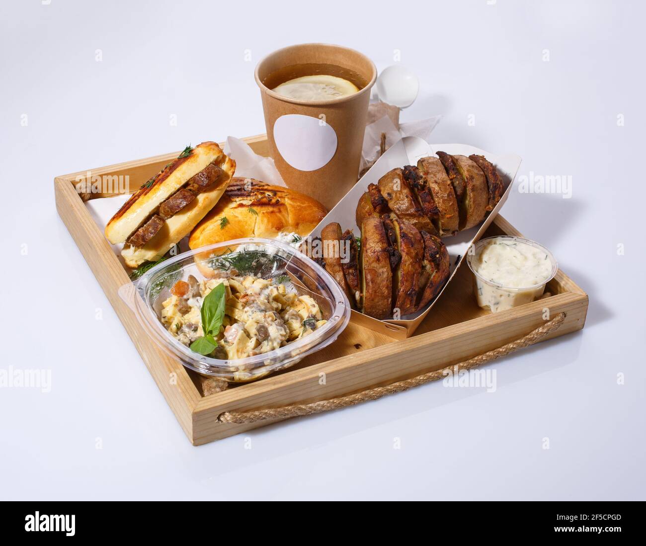 Delicious lunch with disposable tableware on wooden tray. Lemon tea, salad, baked potatoes with meat, sauce and buns. Stock Photo