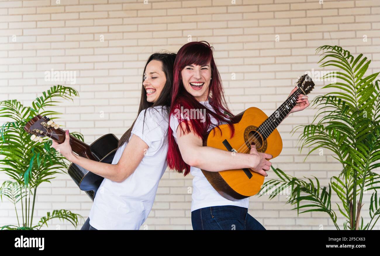 Side view of young women smiling and playing Spanish guitars while standing back to back against brick wall Stock Photo