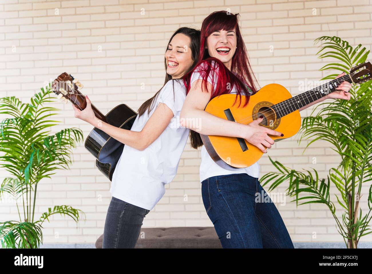 Side view of young women smiling and playing Spanish guitars while standing back to back against brick wall Stock Photo