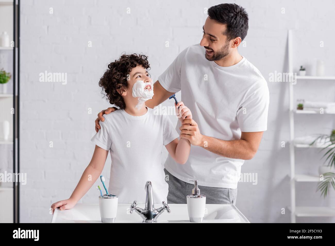 Muslim boy with shaving foam on face holding razor and looking at ...