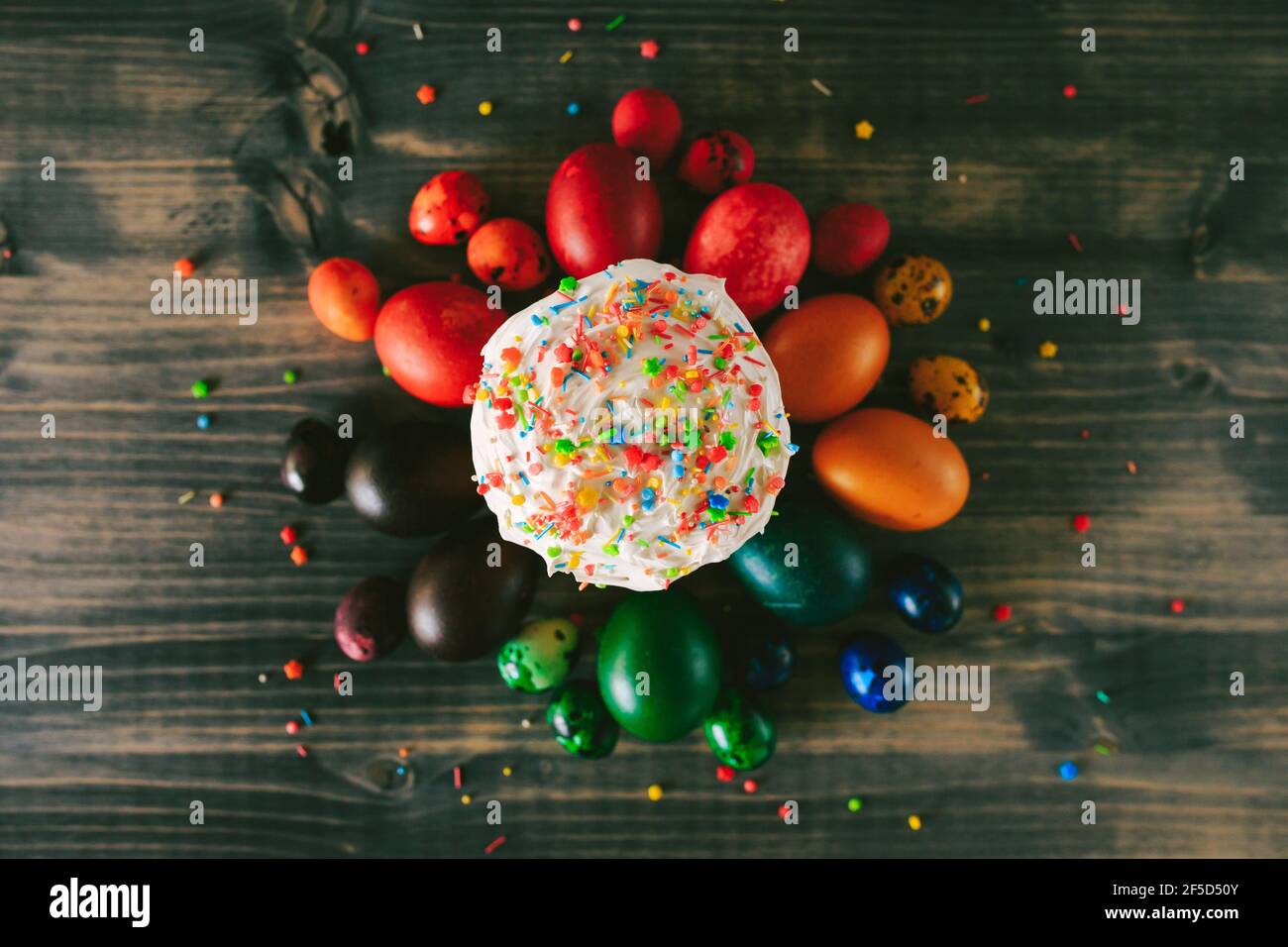 Easter cake sprinkled with multi-colored powder, with multi-colored painted chicken and Easter eggs on a wooden table. Stock Photo
