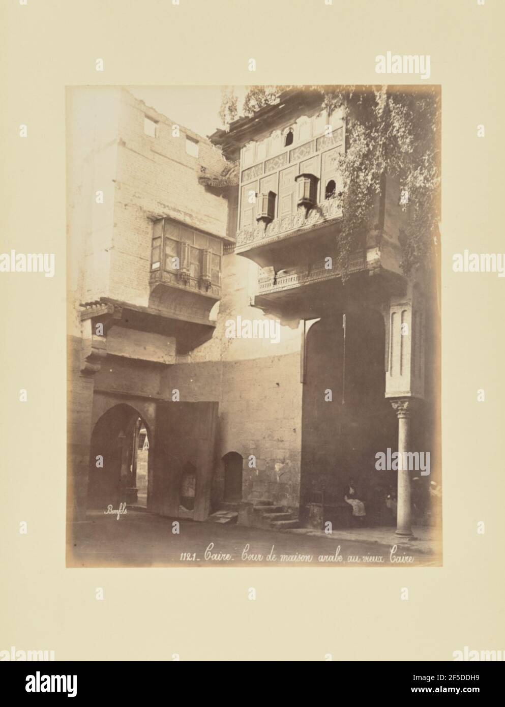 Caire - Cour de maison arabe, au vieux Caire. View of a courtyard surrounded by stone buildings with meshrebeeyehs, a type of oriel windows enclosed with carved wood latticework. A section of the building on the right area of the image appears to be supported by a single decorated pillar, under which a number of people are sitting.. (Recto, print) lower right, inscribed in the negative: '1121   Caire   Cour de maison arabe, au vieux Caire'; Stock Photo