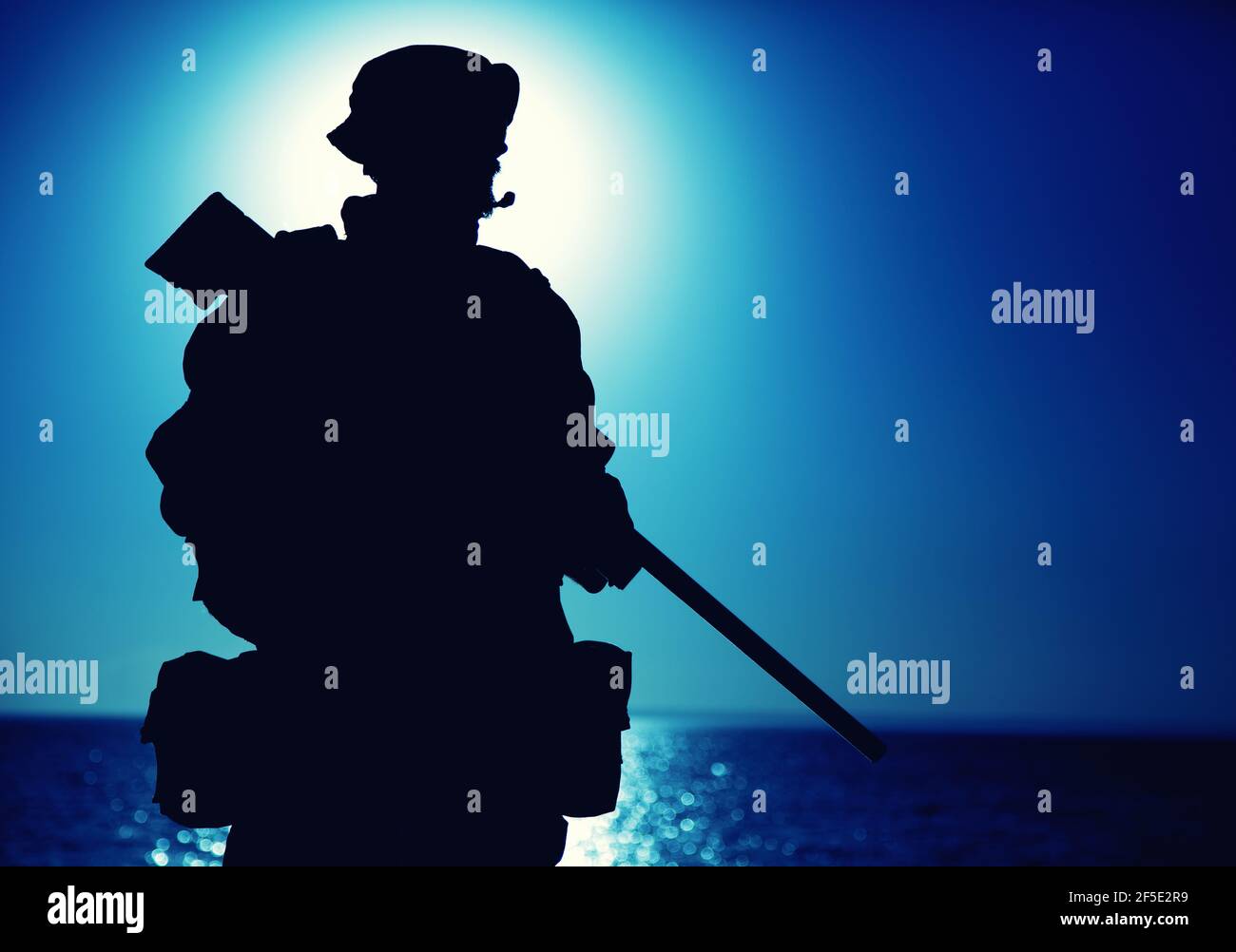 Silhouette of army elite forces fighter standing with sniper rifle on background of blue sky with moon or sun. Sniper or marksman in boonie hat, carrying backpack on mission, patrolling at night Stock Photo