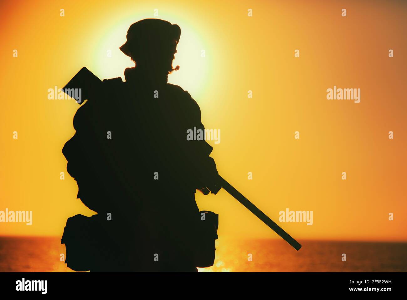 Silhouette of army elite forces sniper, commando shooter in boonie hat, carrying backpack, equipped tactical radio, walking on background of setting or raising sun. Counter terrorist forces soldier Stock Photo