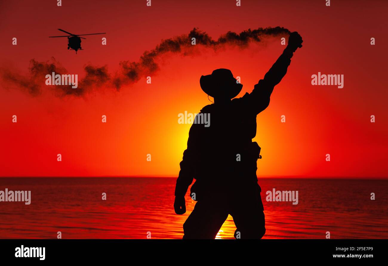 Silhouette of army special operations forces soldier, commando fighter signaling, marking landing spot or evacuation area for helicopter pilot with smoke flair while standing on shore during sunset Stock Photo