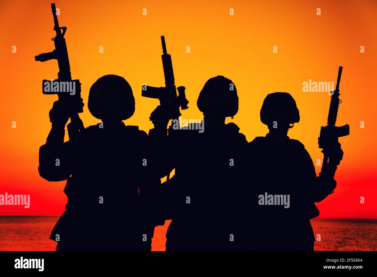 Silhouette of army special operations forces soldiers team, group of Marines or coast guard fighters crew in ammunition and combat helmets standing on seashore at sunset with raised assault rifles Stock Photo