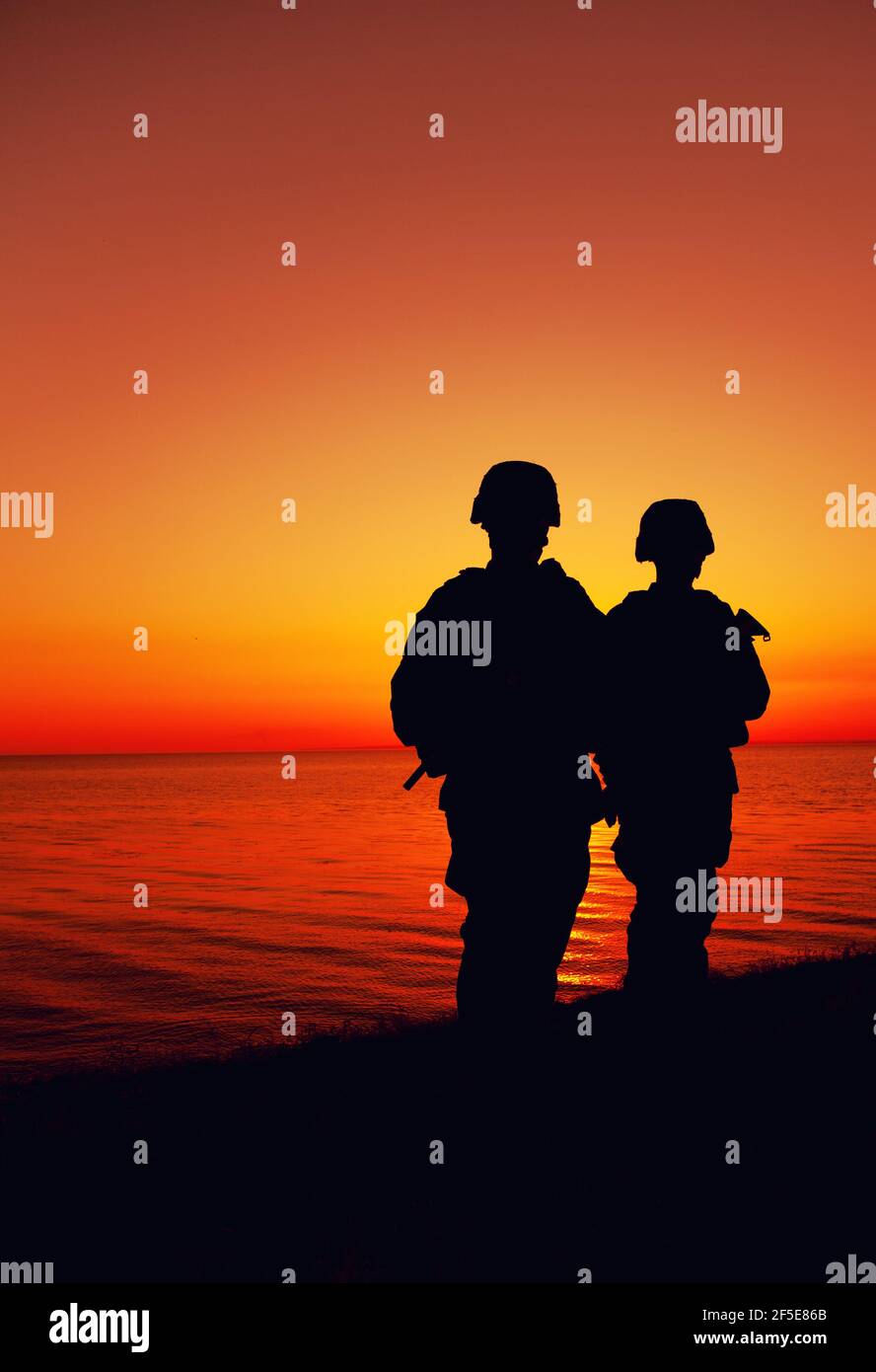 Silhouette of two Marines soldiers, special operations infantrymen or coast guard fighters in uniform and combat helmet, armed service rifles, standing on seashore, patrolling beach at sunset or dawn Stock Photo