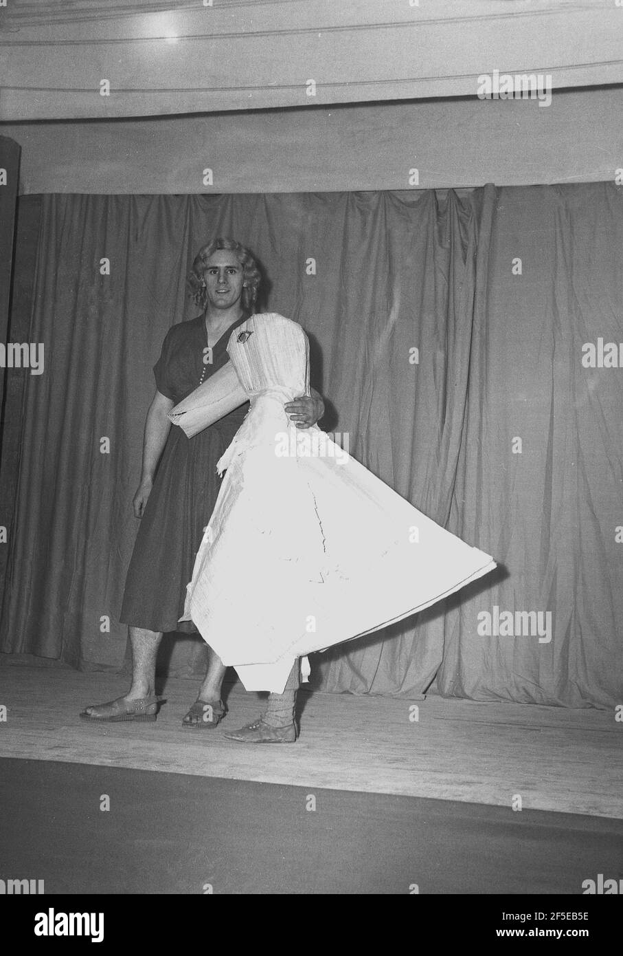 1954, historical, on a stage infront of the curtain wearing costumes, a  tall male actor in a woman's dress and wig and the 'mother goose', performing in the pantomime, 'Mother Goose', England, UK. Originally the imaginary author of a collection of French fairy tales and later of English nursery rhymes, it is said that the character first appeared in 1806 as the performance, 'Harlequin and Mother Goose or The Golden Egg'. Different pantomime adaptions of the Mother Goose story continued throughout the 19th and 20th centuries and it has remained a popular family entertainment show. Stock Photo