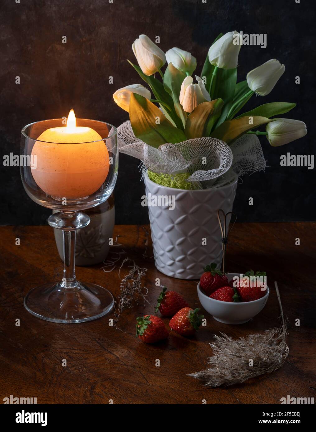 A round candle is lit in a glass candle holder near a vase of white