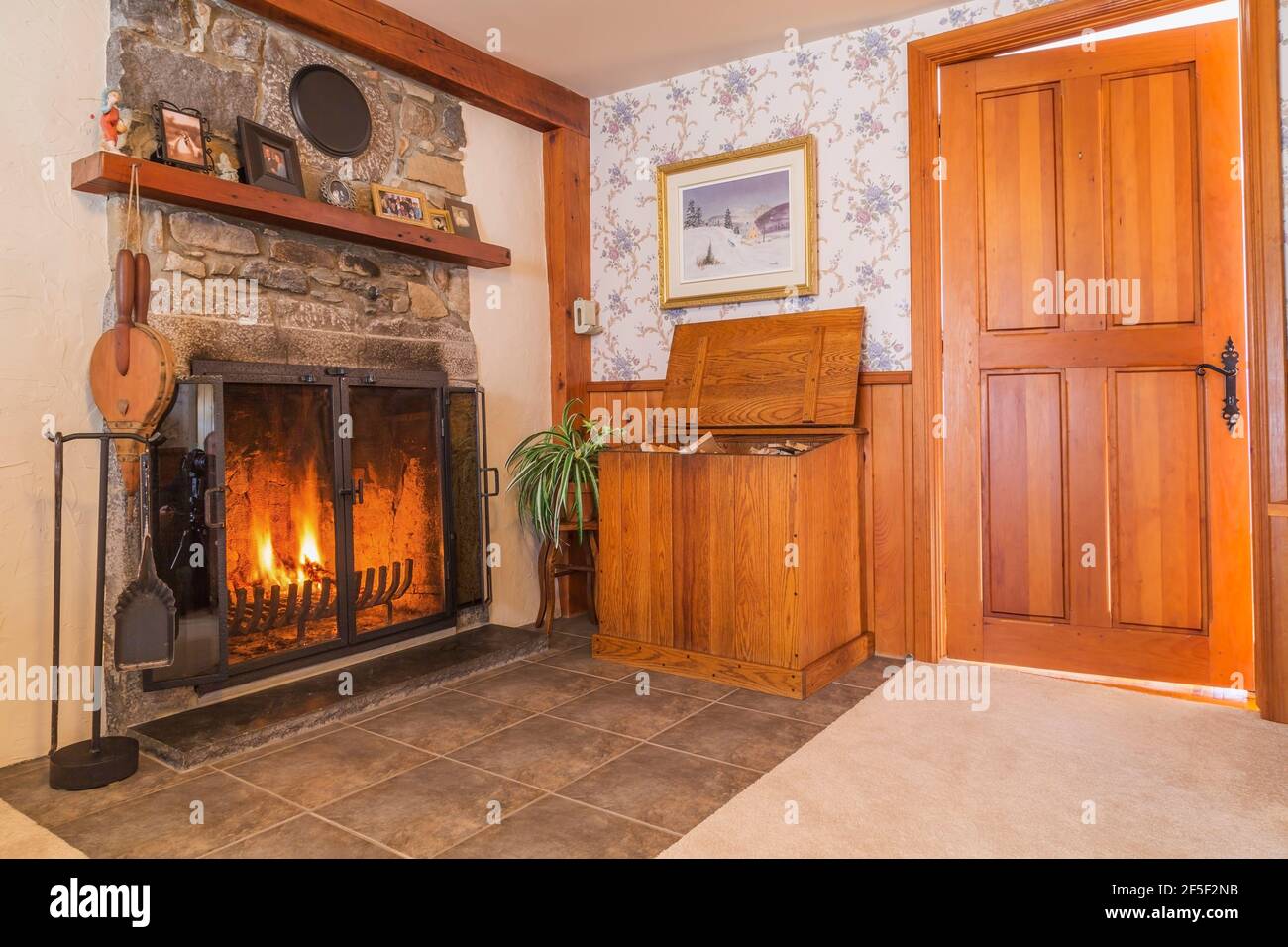 Lit fireplace with fieldstone mantle-piece and firewood storage box in living room with beige carpeting and ceramic tile floor inside an old 1807 home Stock Photo