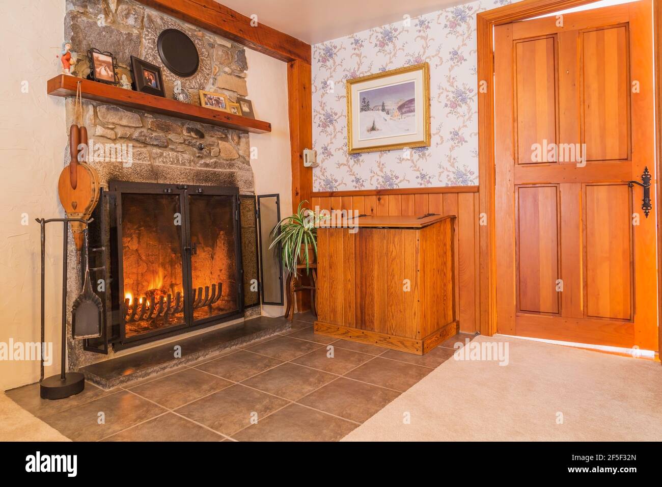 Lit fireplace with fieldstone mantle-piece and firewood storage box in living room with beige carpeting and ceramic tile floor inside an old 1807 home Stock Photo