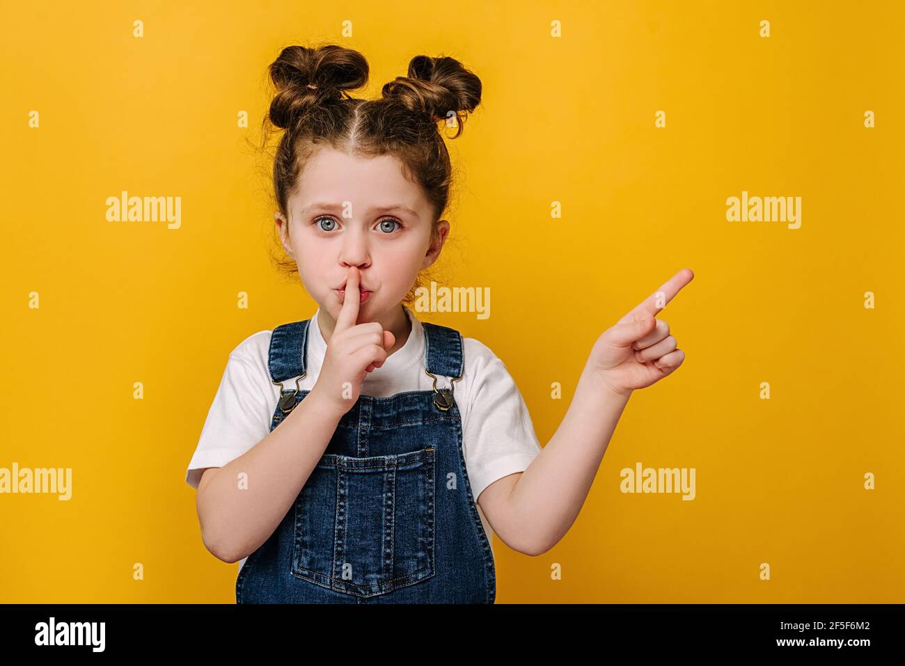 Adorable little girl holding finger on lips symbol of hush gesture of asking to be quiet, isolated over yellow studio background with copy free space Stock Photo