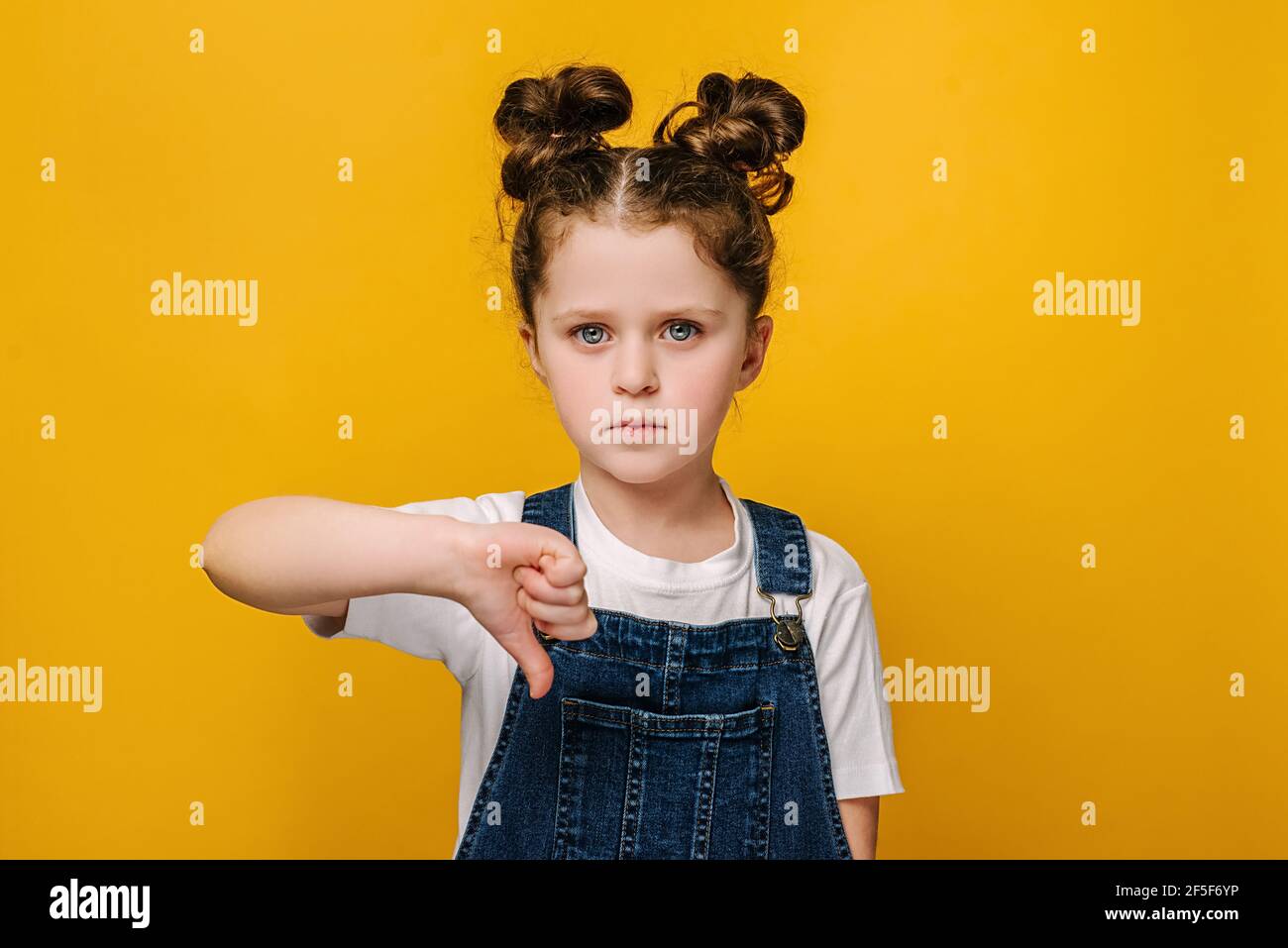 Portrait of serious little girl look at camera show thumb down gesture symbol of negative feedback and dislike, isolated over yellow studio wall Stock Photo