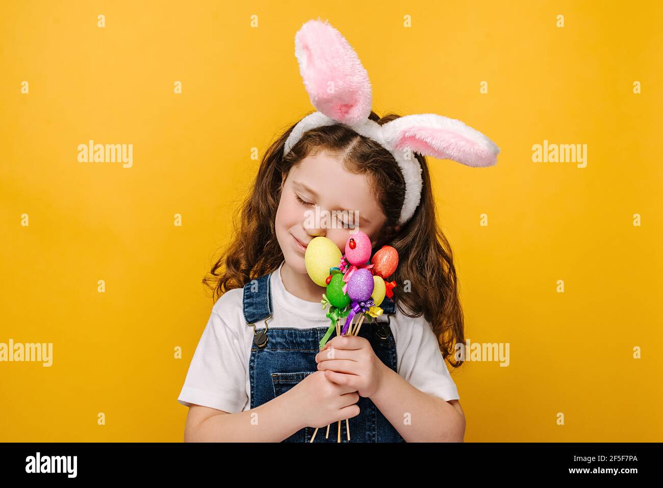 Portrait of dreamy carefree pretty charming preschool girl kid with closed eyes holds small colored eggs on sticks, wears pink bunny fluffy ears Stock Photo