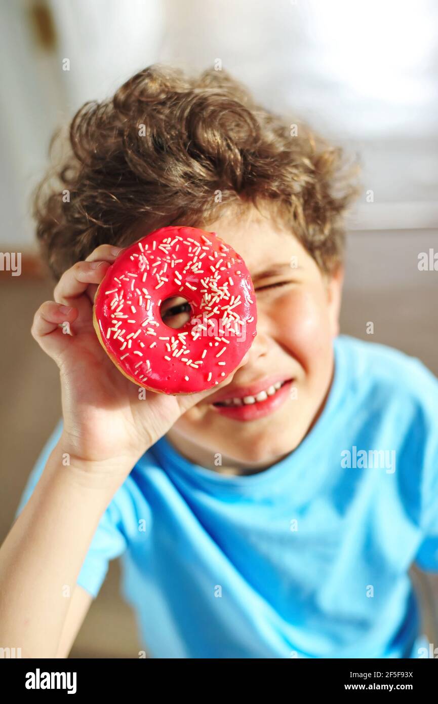 Little happy cute boy looks through a donut like glasses. Delicious food for children. Have fun at home with sweet food. Stock Photo