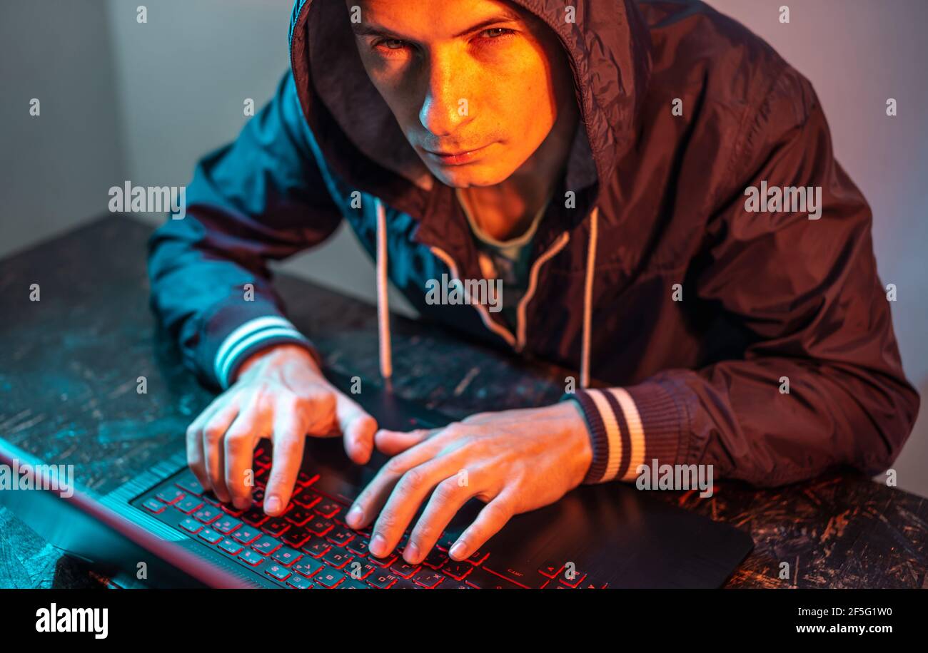 A hooded hacker is typing on a laptop keyboard in a dark room under a neon light. The concept of cybercrime fraud and identity theft Stock Photo