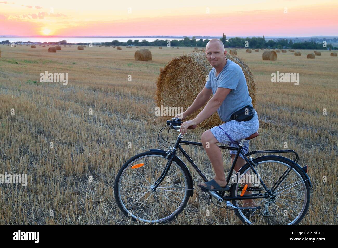 An adult man on a bicycle on a mowed field next to a straw walker at sunset. Sports and holiday activities in the countryside. Stock Photo