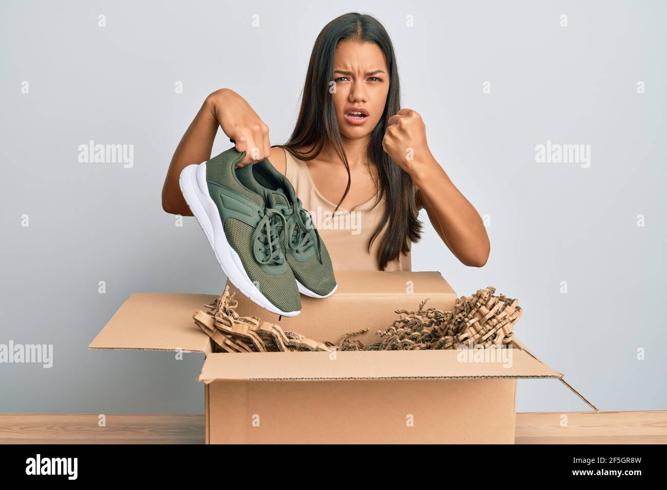 Beautiful hispanic woman taking sneaker shoes from box annoyed and frustrated shouting with anger, yelling crazy with anger and hand raised Stock Photo