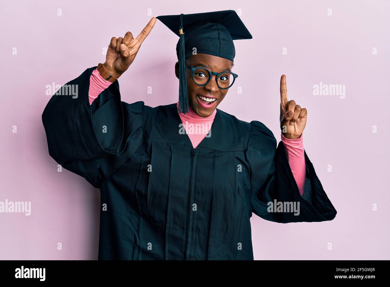 Young african american girl wearing graduation cap and ceremony robe smiling amazed and surprised and pointing up with fingers and raised arms. Stock Photo