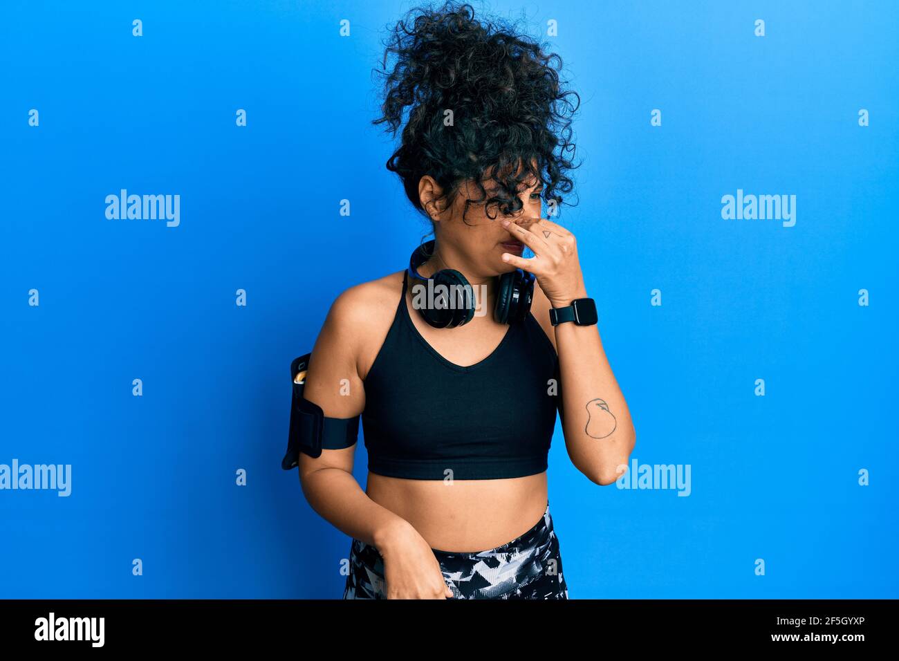 Young hispanic woman wearing gym clothes and using headphones smelling