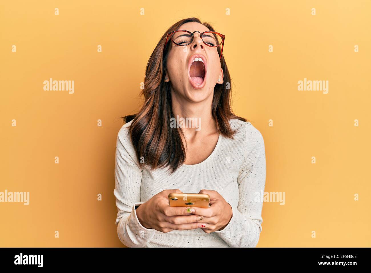 Young brunette woman using smartphone over yellow background angry and ...