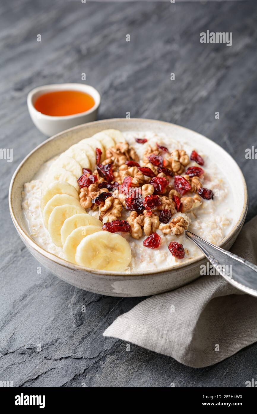 Healthy cereal breakfast, oatmeal topped with banana slices, dried cranberries, walnut, served with honey in a bowl Stock Photo