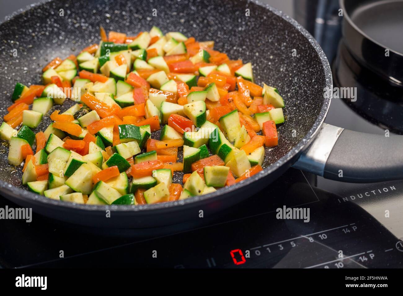 Diced carrots, paprika, and zucchini are sautéed in a black, non-stick frying pan on a modern induction stove Stock Photo