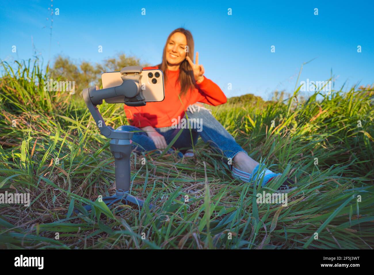 Woman vlogger sitting outside on the grass and live streaming with phone and gimbal. Taking pictures and live video. Blogging. Stock Photo