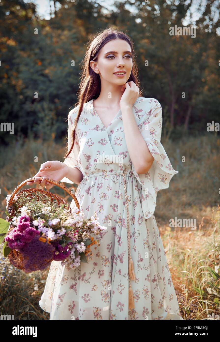 Beautiful smiling young girl in dress holds basket with flowers in hand. Outdoor portrait at meadow Stock Photo