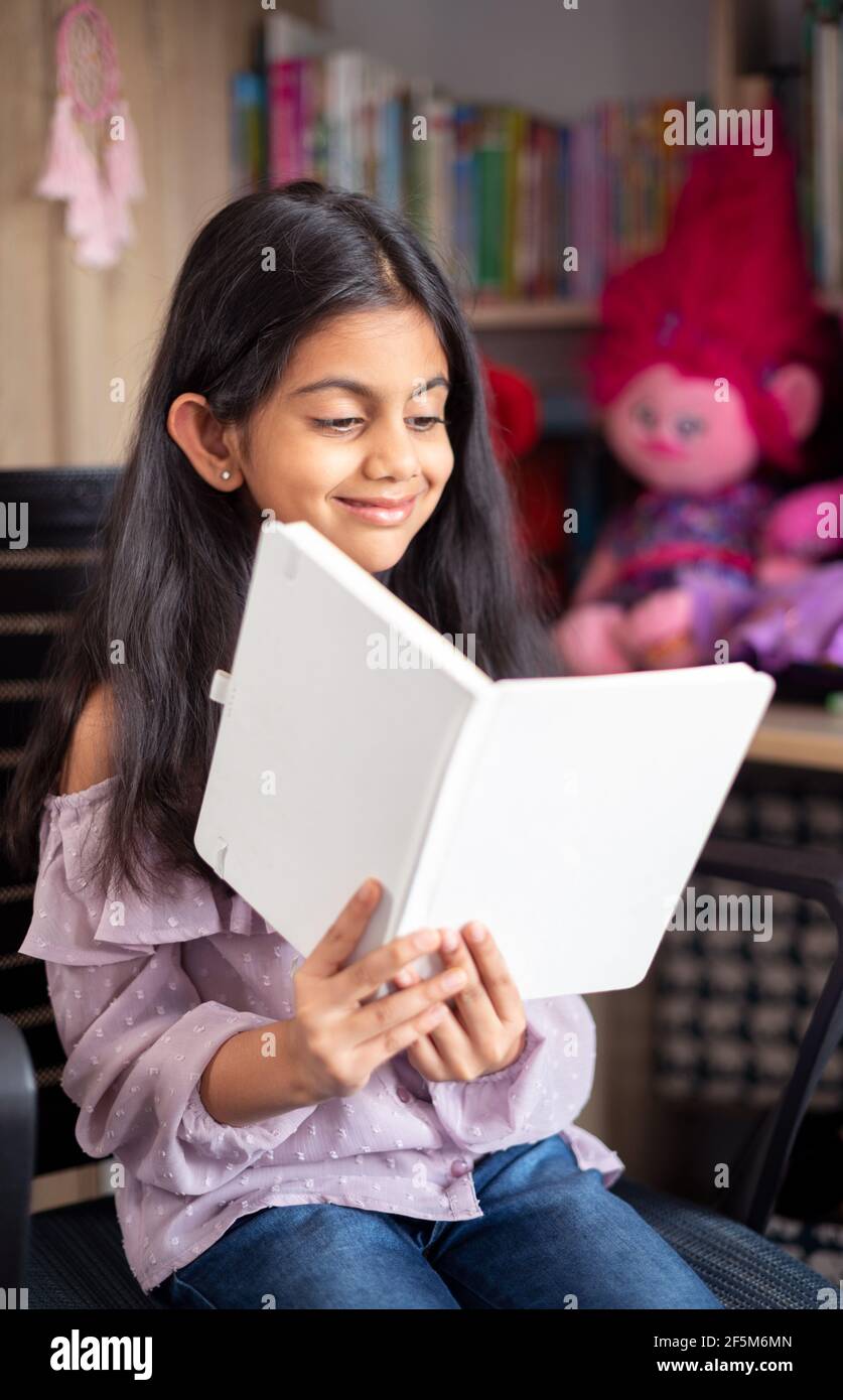 Portrait of a cute Indian school girl sitting on a chair, smiling and reading a diary book surrounded by toys Stock Photo